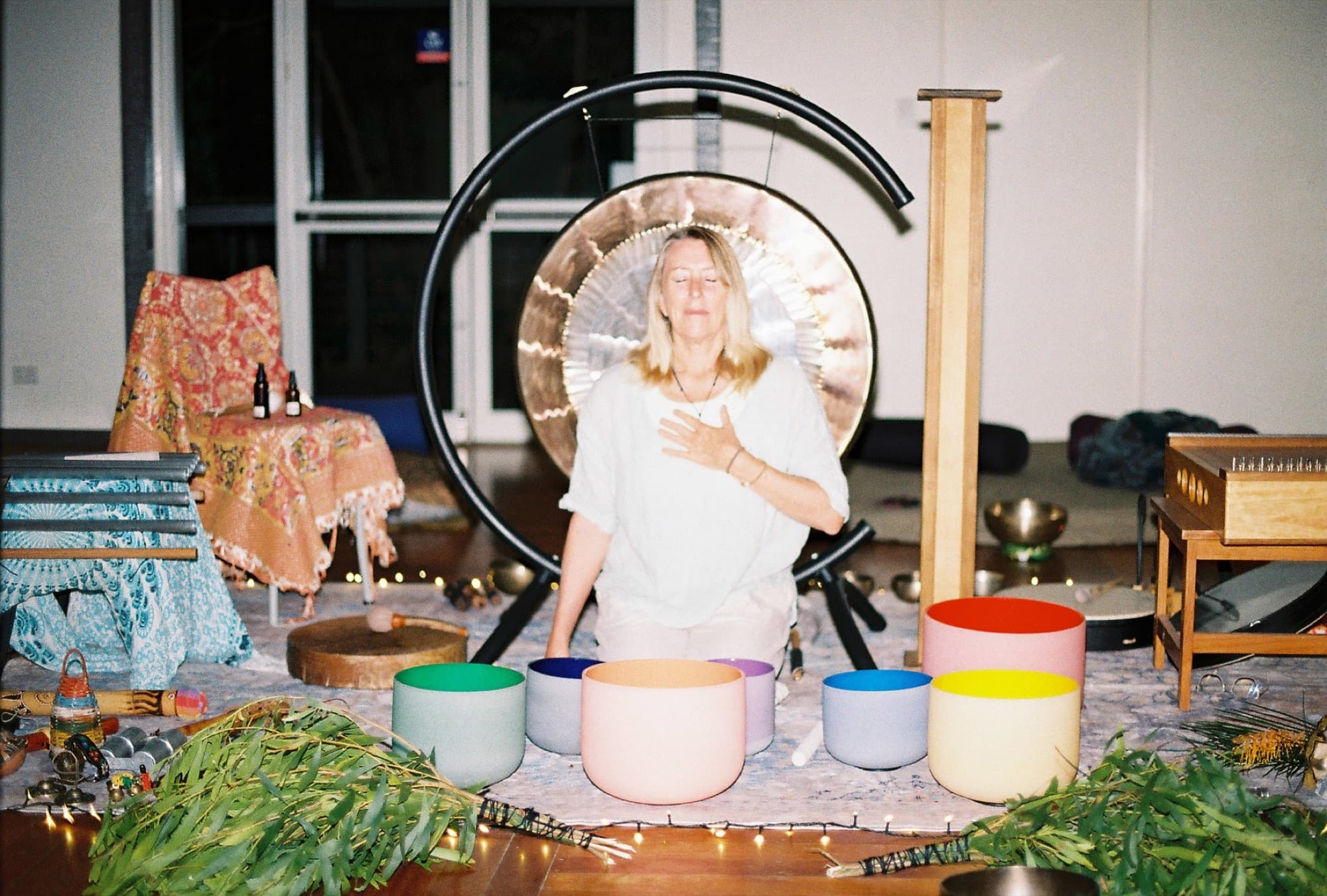 Justine seated in her studio, hand on heart, with gong, crystal bowls, and fairy lights